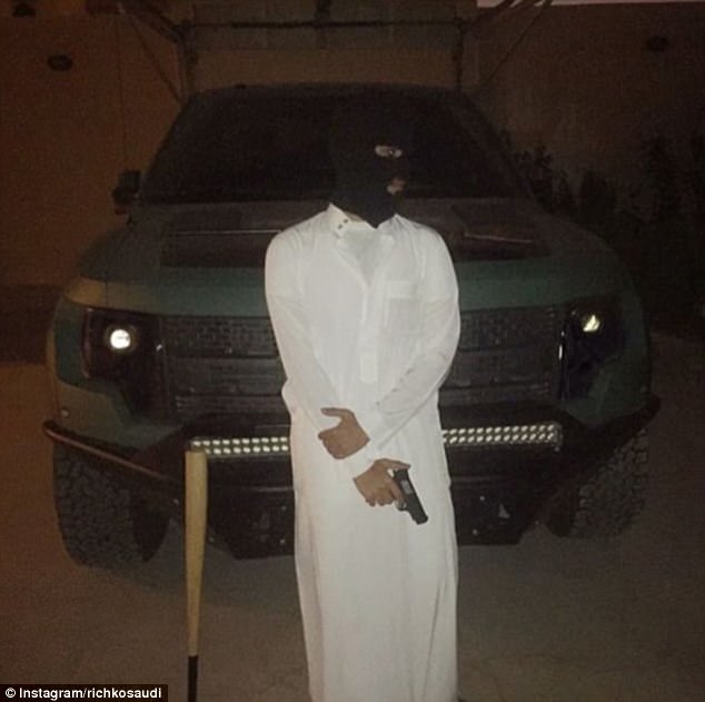 Protection! A man in a balaclava poses in front of a luxury vehicle brandishing a handgun, with a baseball bat propped up nearby